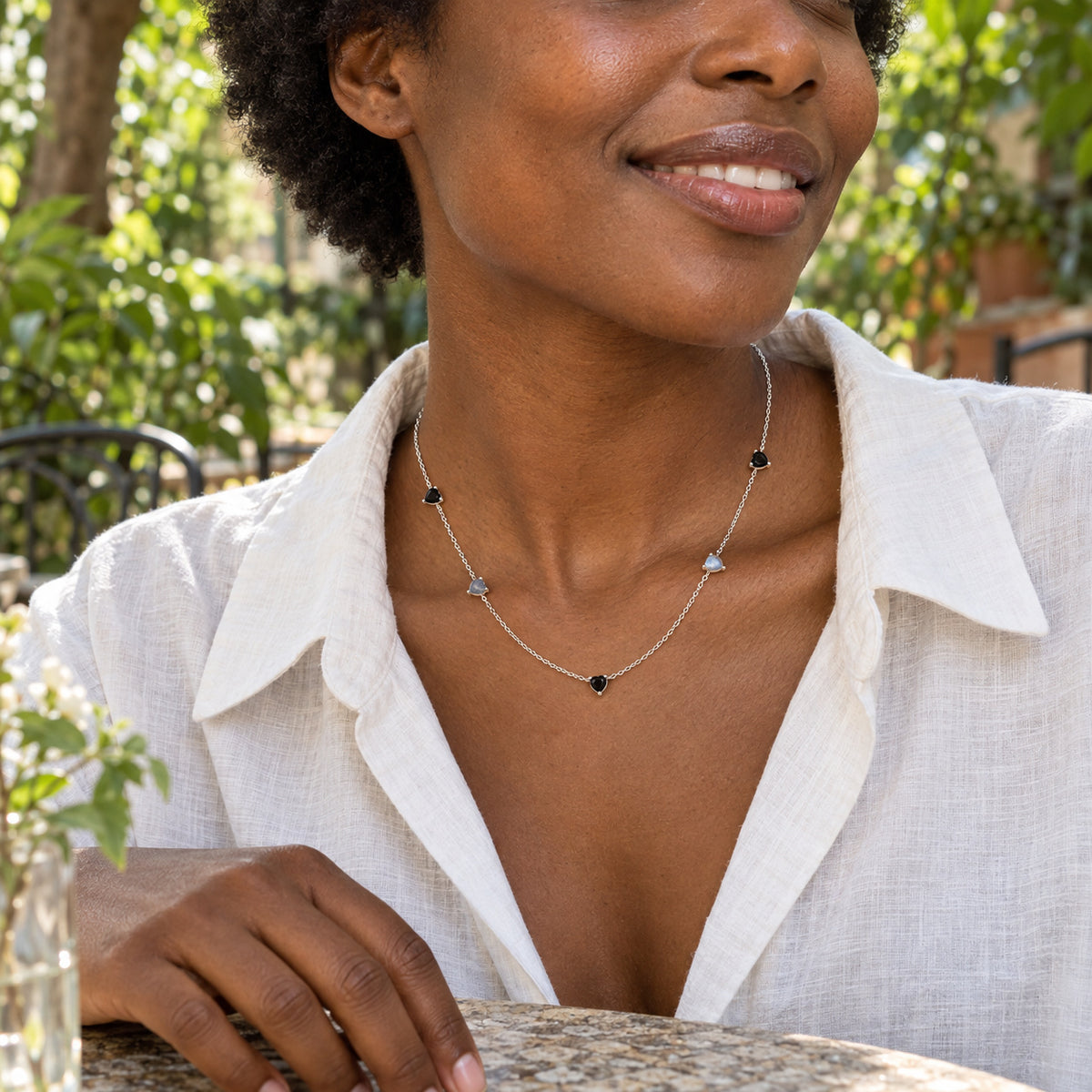 A woman in a white linen shirt wears the silver five-heart necklace at a leafy outdoor cafe.