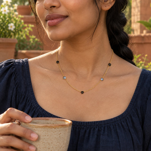 A woman in a navy top wears the yellow gold heart-station necklace while holding a ceramic cup on a garden terrace.