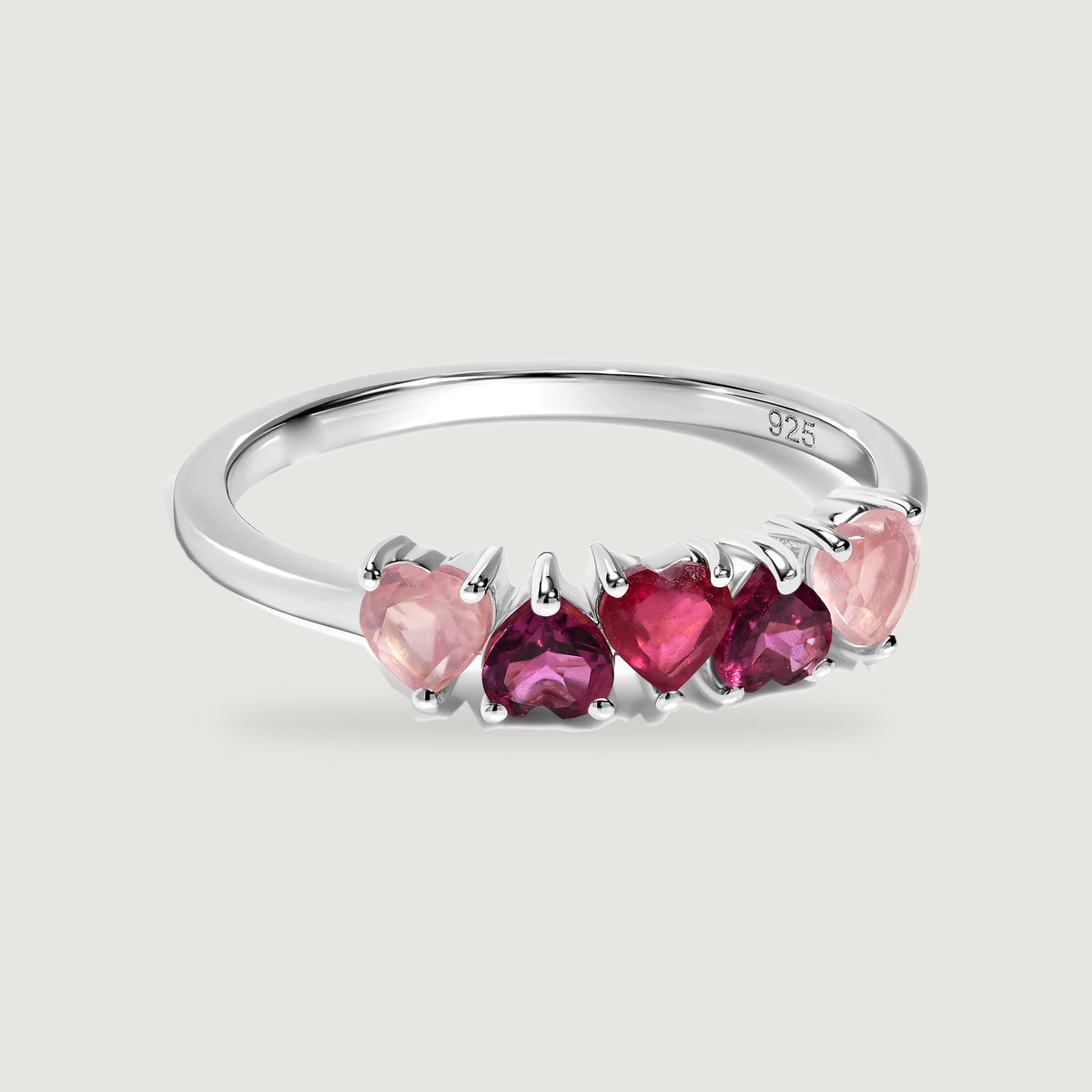 Five heart-shaped pink and raspberry stones sit across a polished silver ring against a soft gray background.