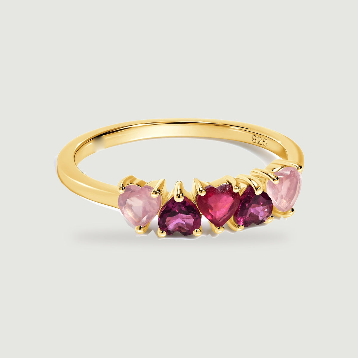A yellow gold ring with five alternating pink and raspberry heart-shaped stones is centered on a pale gray surface.