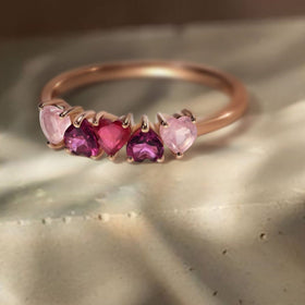 The rose gold heart-stone ring rests on a sunlit stone tabletop with soft diagonal shadows.