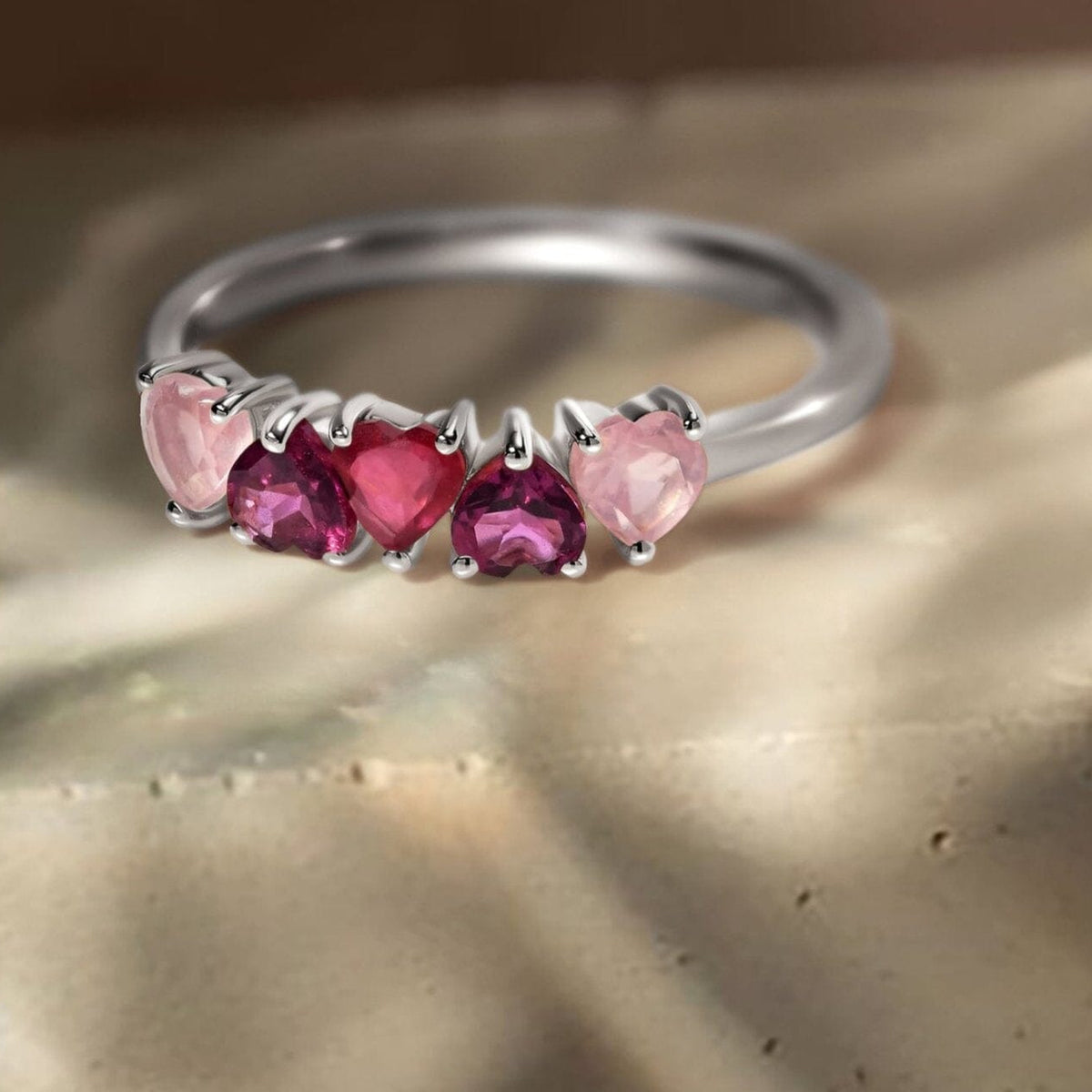 A silver ring with five pink and raspberry heart-shaped stones lies on a textured stone surface in natural light.