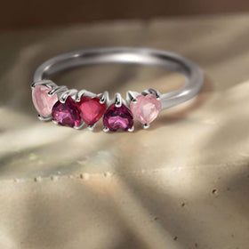 A silver ring with five pink and raspberry heart-shaped stones lies on a textured stone surface in natural light.