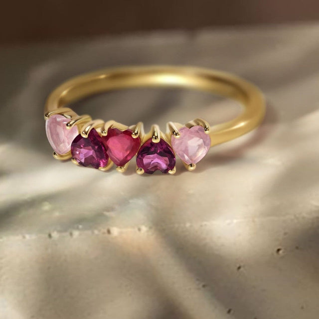 The yellow gold ring rests on a warm stone tabletop with its row of five heart-shaped stones facing forward.