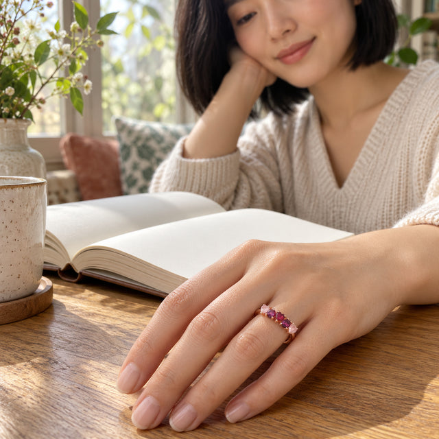 A rose gold heart-stone ring is worn on a hand resting on a wooden table beside an open book.