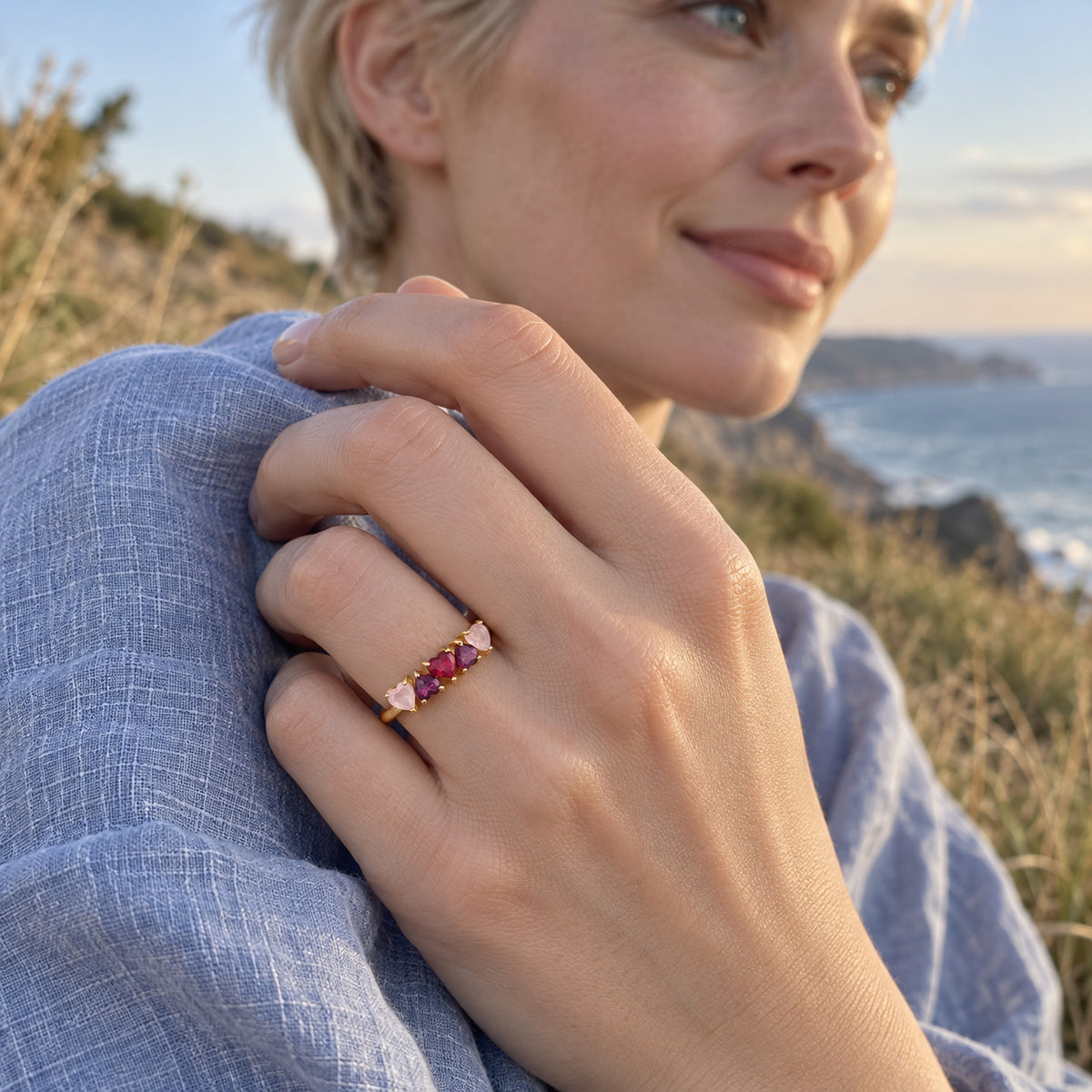 A yellow gold heart-stone ring is shown on a hand touching a blue linen sleeve beside the coast.