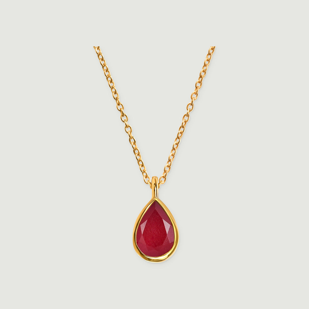 A yellow gold necklace with a faceted deep pink teardrop pendant is centered on a light gray background.
