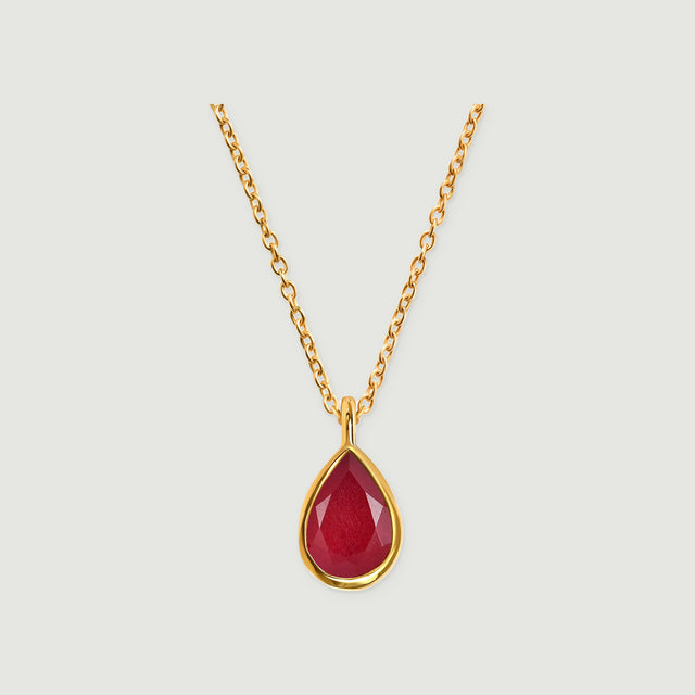 A yellow gold necklace with a faceted deep pink teardrop pendant is centered on a light gray background.