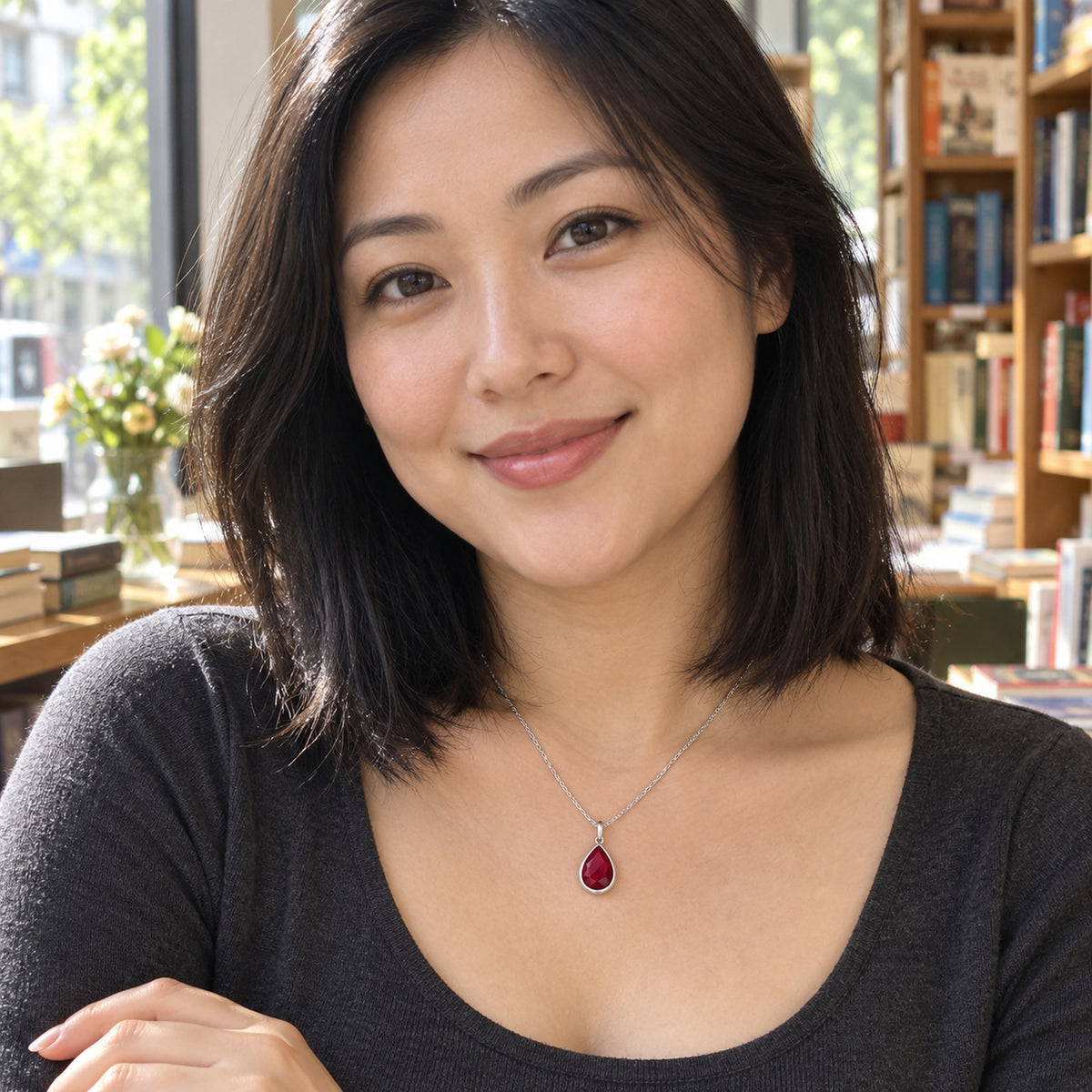 A woman with straight black hair wears a silver pink gemstone pendant and a charcoal top beside a bookshop window.
