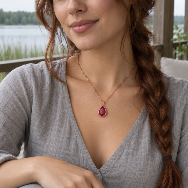 A woman with an auburn side braid wears a rose gold pink teardrop necklace and a gray blouse on a lakeside porch.