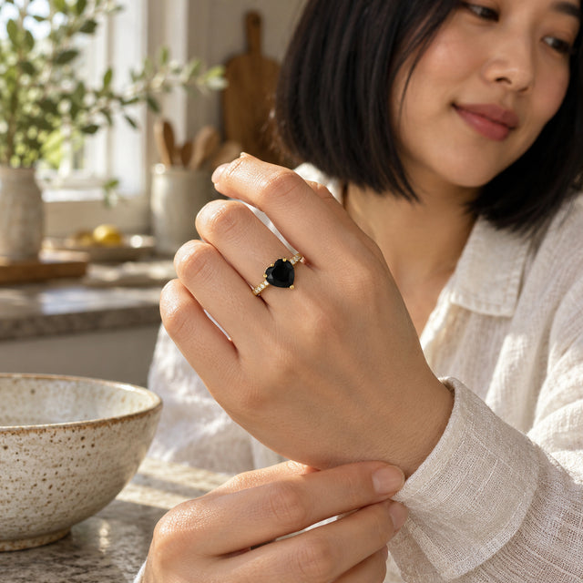 A woman wears a yellow gold vermeil black heart ring while adjusting a linen sleeve in a sunlit kitchen.