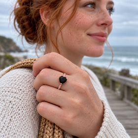 A freckled woman wears a rose gold vermeil black heart ring while holding a woven bag strap beside the sea.