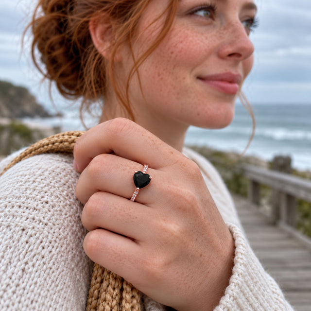A freckled woman wears a rose gold vermeil black heart ring while holding a woven bag strap beside the sea.