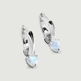Two polished silver-tone hoop earrings hold heart-shaped moonstones on a light gray surface.
