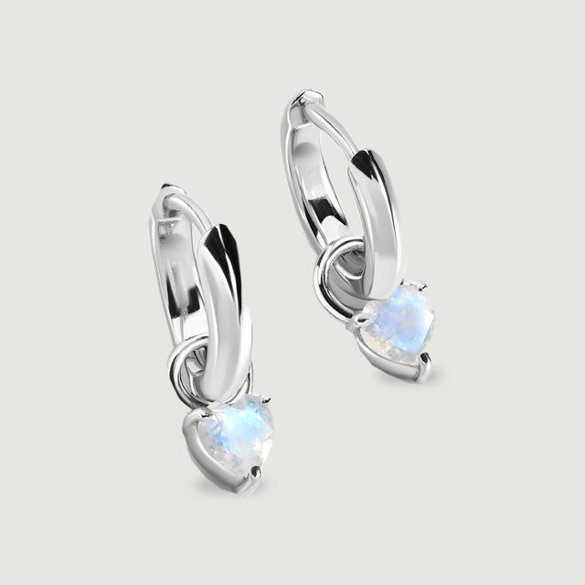 Two polished silver-tone hoop earrings hold heart-shaped moonstones on a light gray surface.