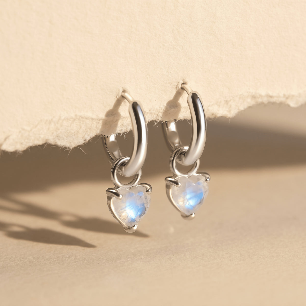 Two silver-tone hoops with blue-flashing heart-shaped stones hang from textured paper above a beige tabletop.