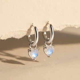 Two silver-tone hoops with blue-flashing heart-shaped stones hang from textured paper above a beige tabletop.