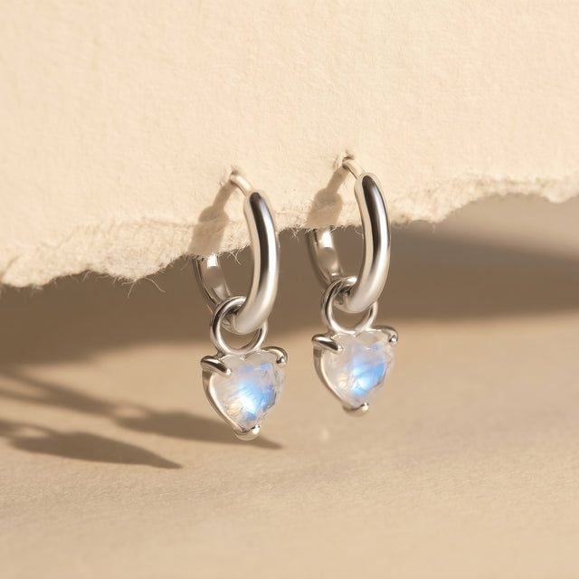 Two silver-tone hoops with blue-flashing heart-shaped stones hang from textured paper above a beige tabletop.