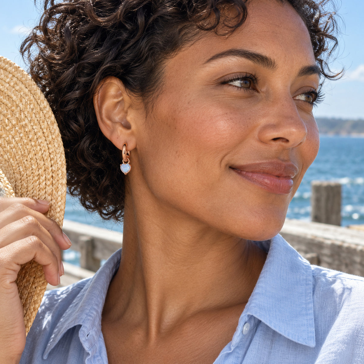 A woman in a light blue shirt wears a rose-gold-tone moonstone hoop beside the sea.