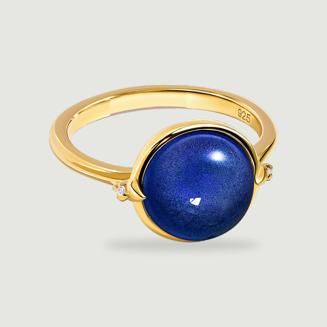 A yellow gold ring with a round blue cabochon and two small clear side stones stands on a pale grey surface.