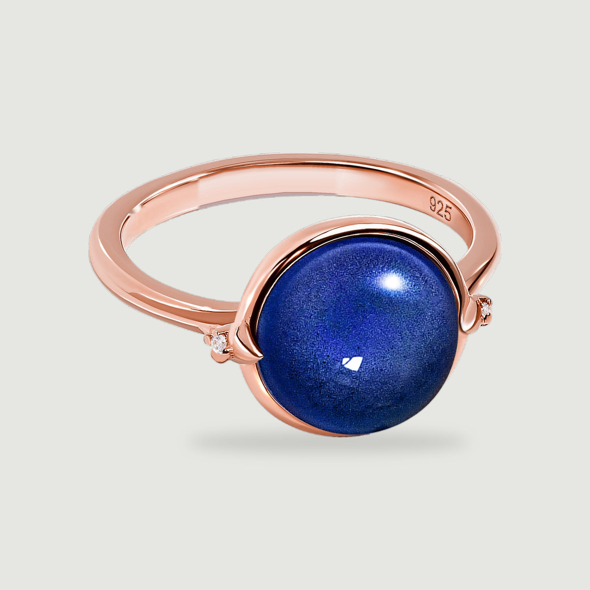 A rose gold ring with a deep blue round stone and tiny clear side accents is shown at an angle on light grey.