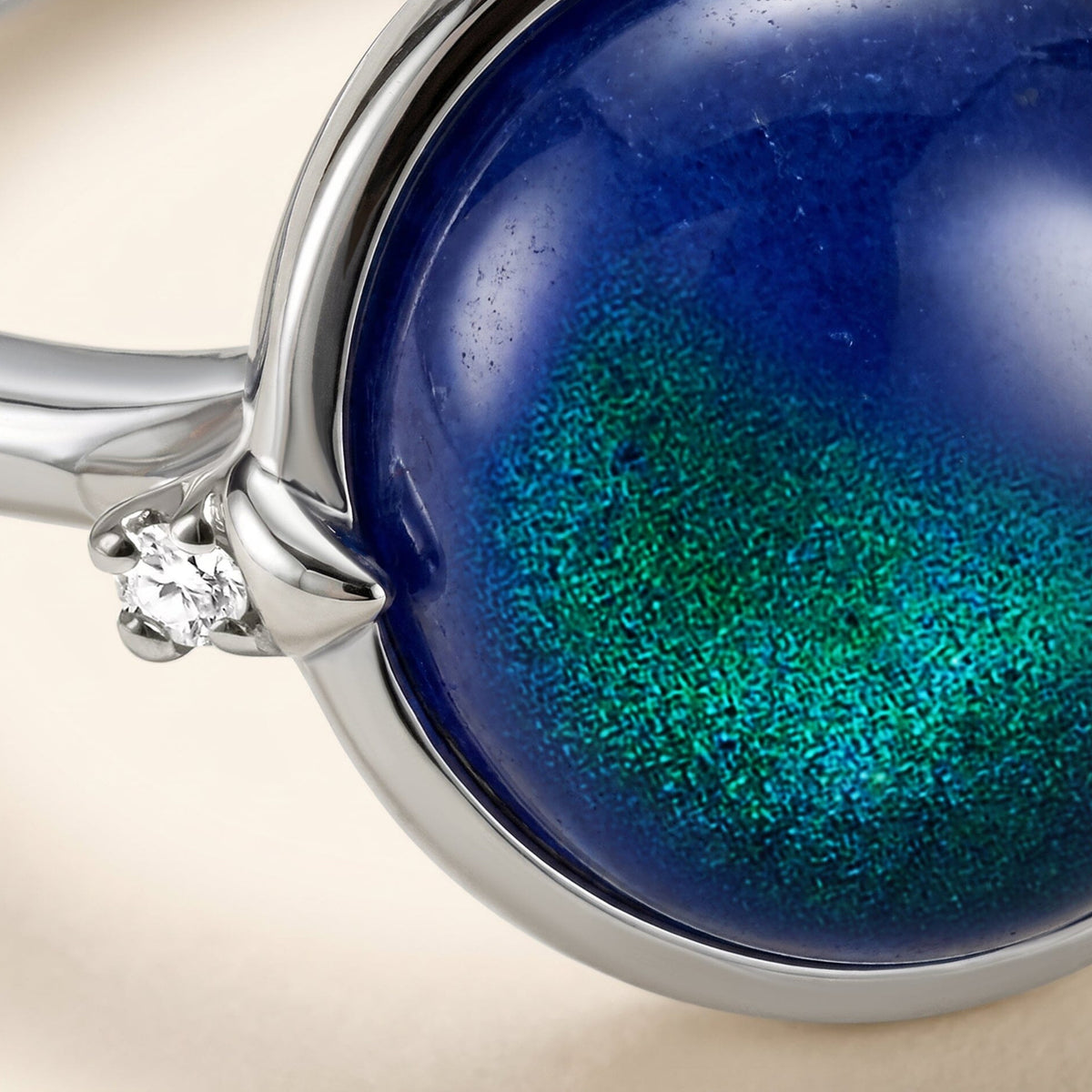 The rounded blue stone and a bright clear side stone fill the frame above a silver band and a pale tabletop.