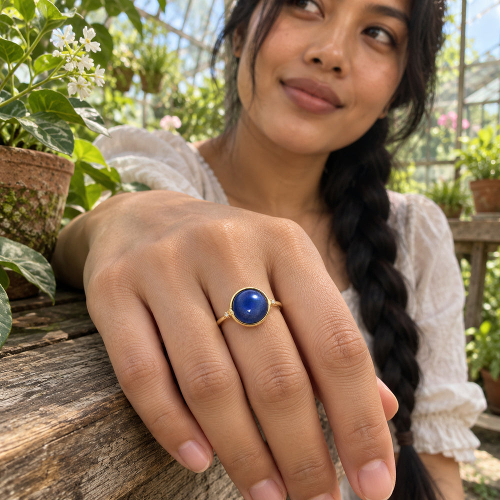 A yellow gold ring with a blue round stone is worn on a hand resting on a wooden bench among greenhouse plants.