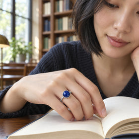 A silver ring with a domed blue stone is worn on a hand resting on an open book beside a library window.