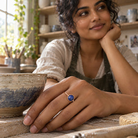 A rose gold ring with a round blue cabochon is worn on a hand beside a glazed bowl in a pottery studio.