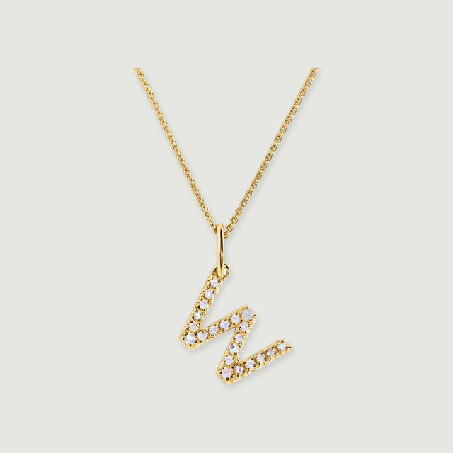 A stone-set W-shaped pendant hangs diagonally from a gold-tone chain on a soft grey surface.