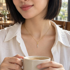 A woman with a black bob wears a gold-tone I pendant above an ivory shirt while holding a ceramic cup in a cafe.