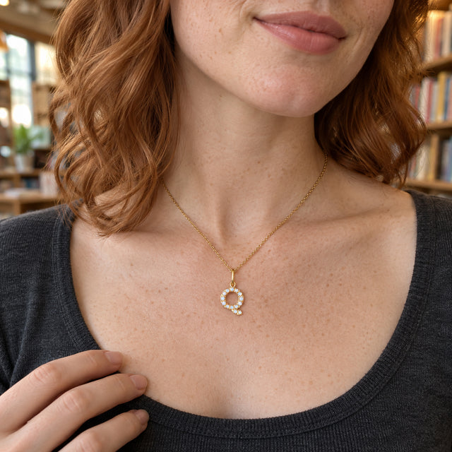 A freckled woman with copper waves wears a pale stone-set Q pendant over a charcoal neckline in a bookshop.