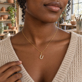 A woman with dark skin and curled hair wears a gold-tone U necklace above an oatmeal sweater in a ceramics studio.