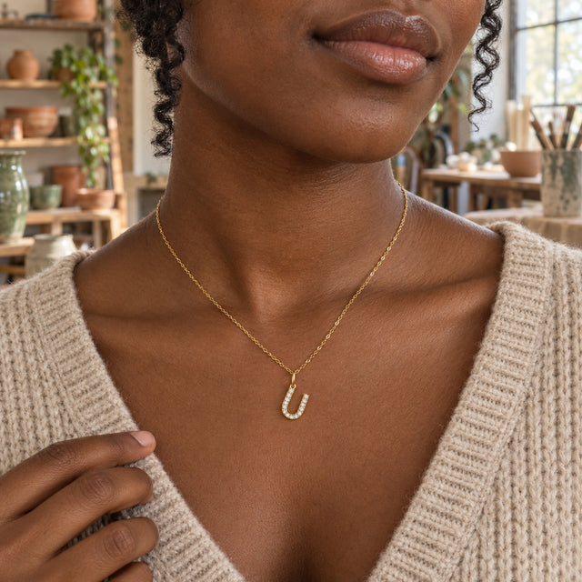 A woman with dark skin and curled hair wears a gold-tone U necklace above an oatmeal sweater in a ceramics studio.