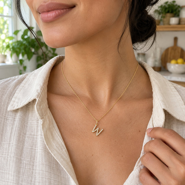 A woman with dark hair tied back wears a stone-set W pendant between the open collars of a linen blouse in a bright kitchen.