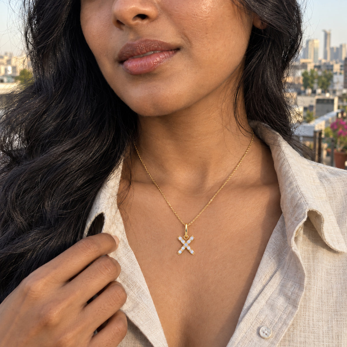 A woman with long dark waves wears a gold-tone X pendant above a beige shirt on a rooftop overlooking city buildings.