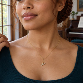 A woman with an auburn braided updo wears a pale stone-set Y necklace over a dark teal neckline beside a window and piano.
