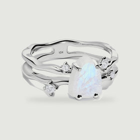 A white gold ring with a pale blue raw moonstone and four round clear accent stones rests on a light gray surface.