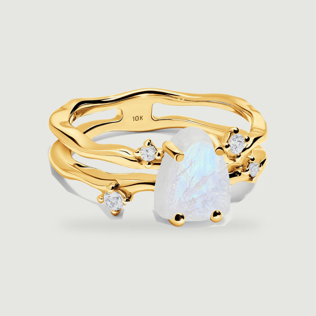A yellow gold ring with three curved bands holds a raw moonstone between four rounded prongs on a light gray surface.