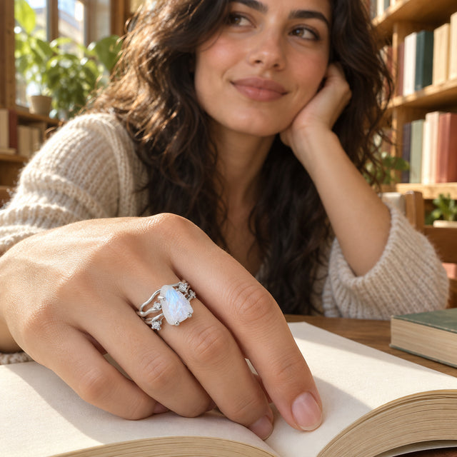 A white gold moonstone ring is worn on a hand resting on an open book in a sunlit bookshop.