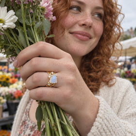 A yellow gold moonstone ring is visible on a freckled hand holding a bouquet at an outdoor market.
