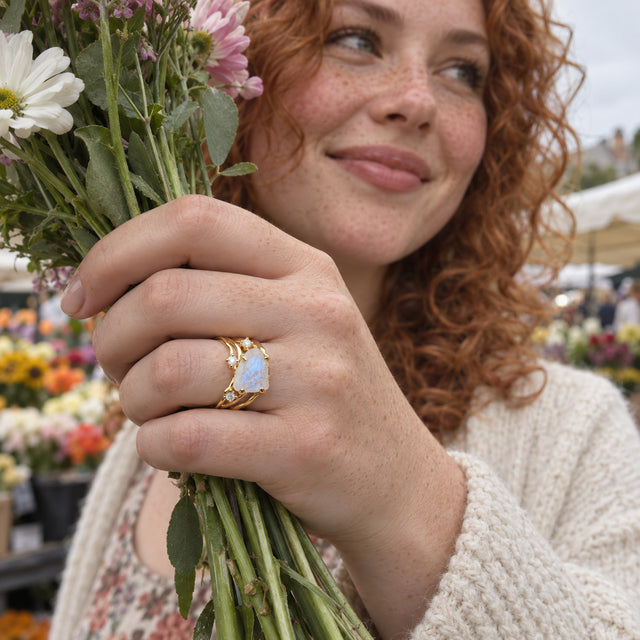 A yellow gold moonstone ring is visible on a freckled hand holding a bouquet at an outdoor market.