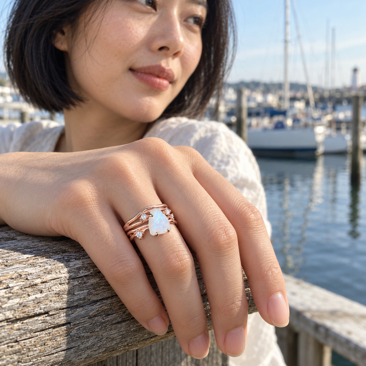 A rose gold moonstone ring is worn on a hand resting on a weathered wooden railing beside a harbor.