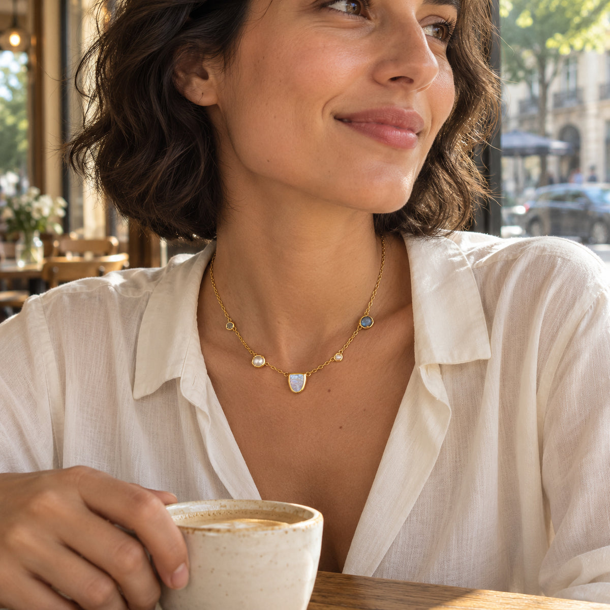 A woman in an ivory blouse wears a gold-tone moonstone and pearl necklace while holding a ceramic cup in a sunlit cafe.
