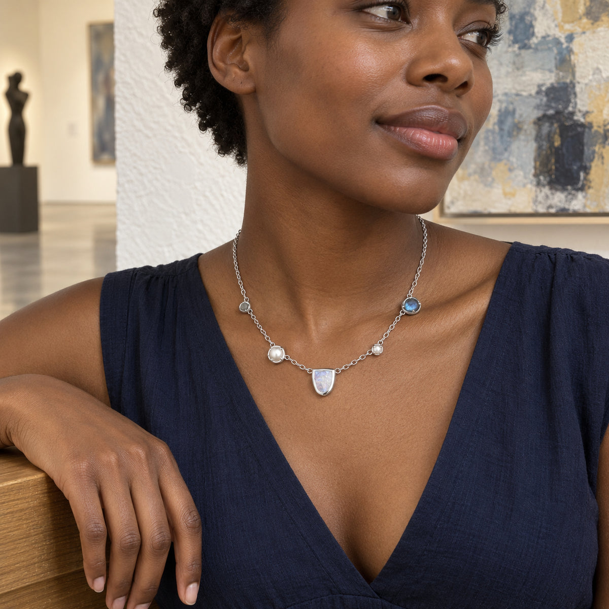 A woman in a navy V-neck dress wears a silver-tone necklace with a shield-shaped moonstone and round pearl accents in an art gallery.