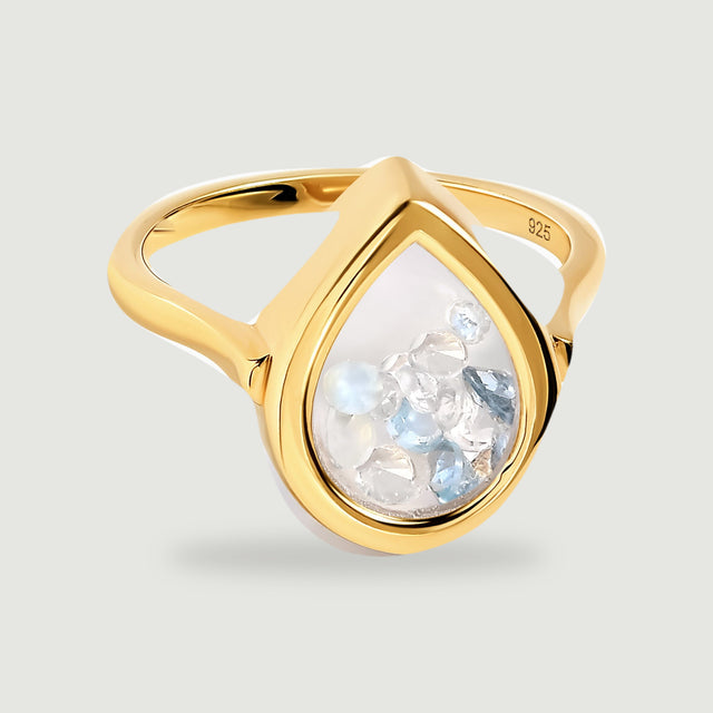 A yellow gold ring with a teardrop glass face rests above a soft shadow on a light grey surface.