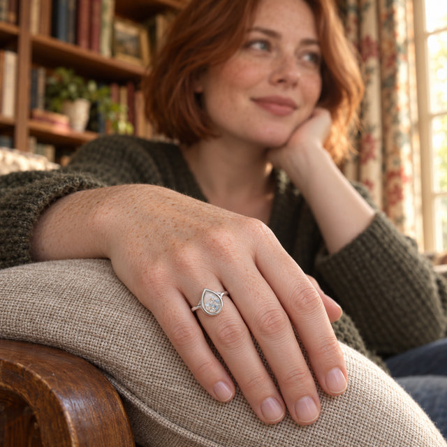 A silver pear-shaped ring filled with tiny blue and clear gems is worn on a freckled hand resting on a linen cushion in a library.