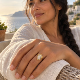 A yellow gold ring with a glass chamber of blue and white gems is worn on a hand beside a linen sleeve on a coastal terrace.