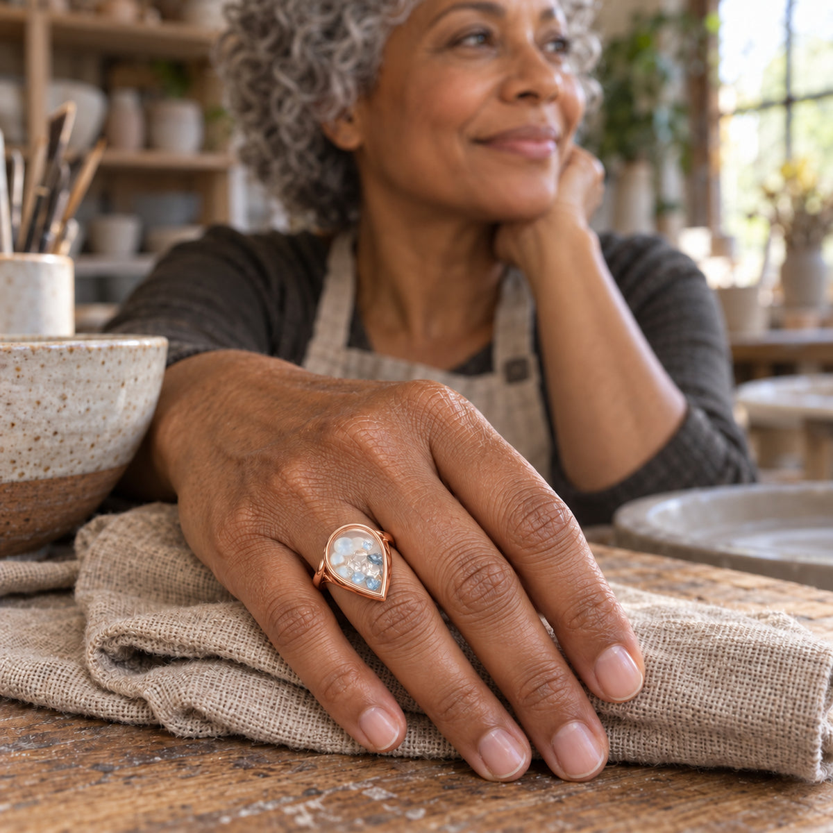 A rose gold teardrop ring containing pale blue and translucent gems is worn on a hand resting on woven cloth in a pottery workshop.
