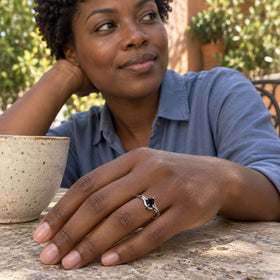 A silver ring with a small black center stone is worn on a hand resting beside a ceramic cup on a stone cafe table.